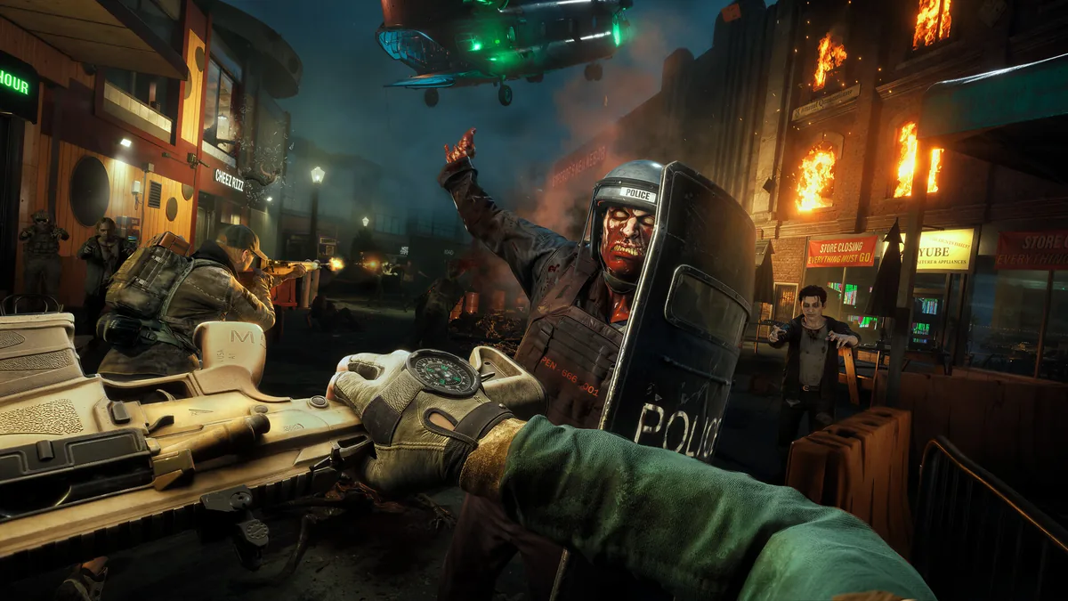 A squad faces a riot-shield zombie on a burning city street in official gameplay.